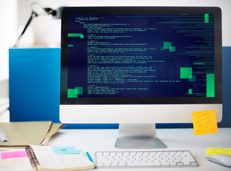 a notebook, keyboard, and a PC monitor with code on the screen